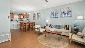 A spacious living room with a wooden floor, a round coffee table, and a sofa with patterned cushions. A dining area with wooden chairs and a table is visible in the background. - Beach Paradise IX-Charming 4BR Townhouse•DOWNTOWN•Short walk to Beach&Boardwalk (Ocean City)
