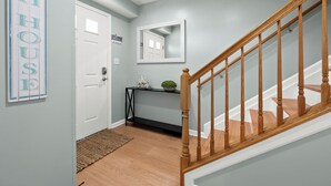 A hallway with a wooden staircase, a white door, a black console table with a plant, and a mirror on the wall. - Beach Paradise IX-Charming 4BR Townhouse•DOWNTOWN•Short walk to Beach&Boardwalk (Ocean City)