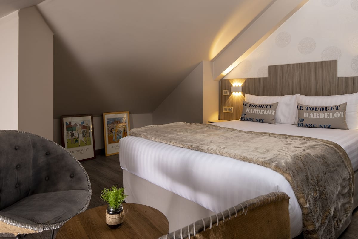 A bedroom with a large bed, a grey armchair, a small table with a plant, and framed pictures on the wall.