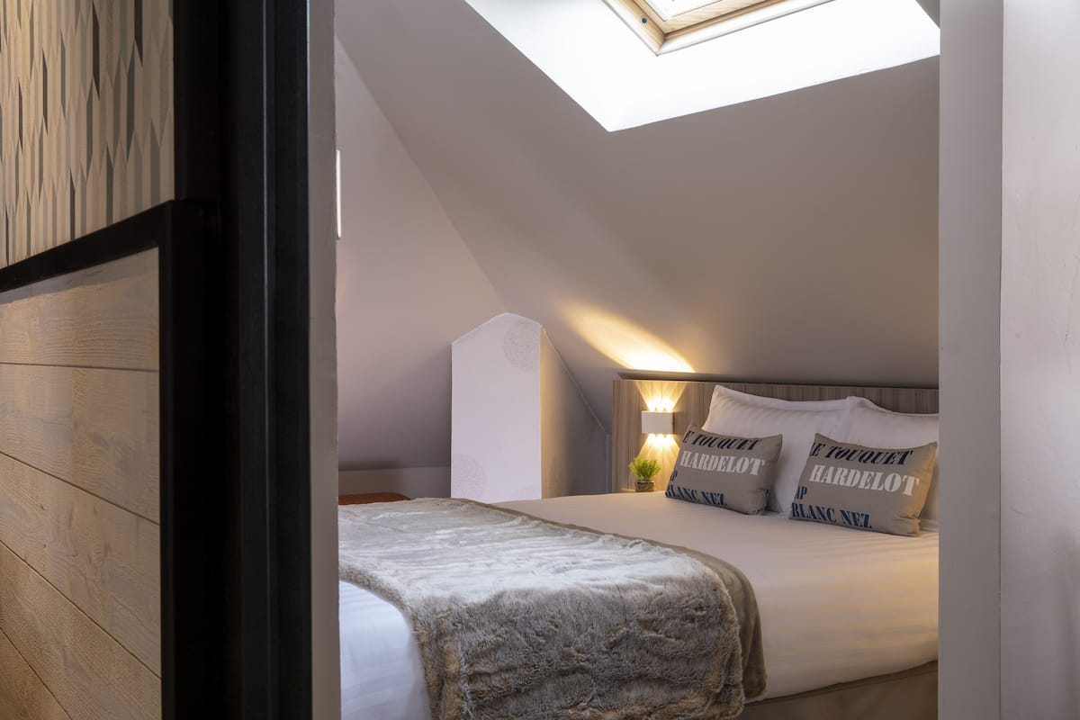 A bedroom with a skylight, a bed with white bedding and patterned pillows, a bedside table with a lamp, and a small plant.