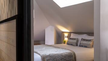 Une chambre avec un puits de lumière, un lit avec une literie blanche et des coussins à motifs, une table de chevet avec une lampe et une petite plante.