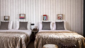 A room with two beds, each with a wooden headboard, a small bedside table with a lamp, and a wall-mounted shelf with books.