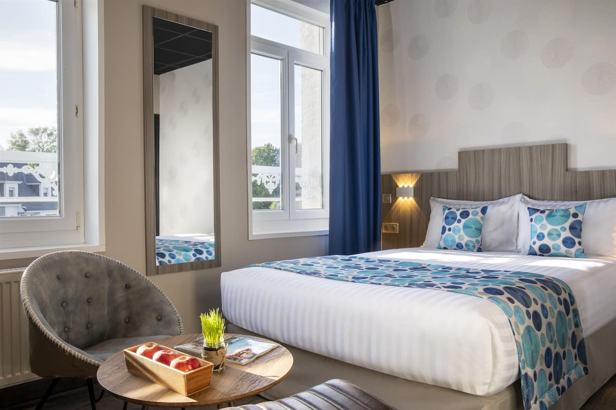 A hotel room with a bed, a chair, a round table with a tray of apples, and a window with a view of buildings.