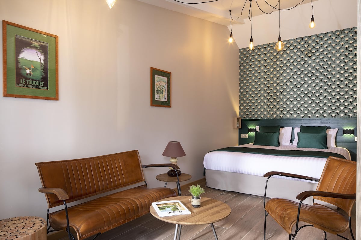 A hotel room with a bed, two brown leather sofas, a small round table with a lamp, and a framed picture on the wall.