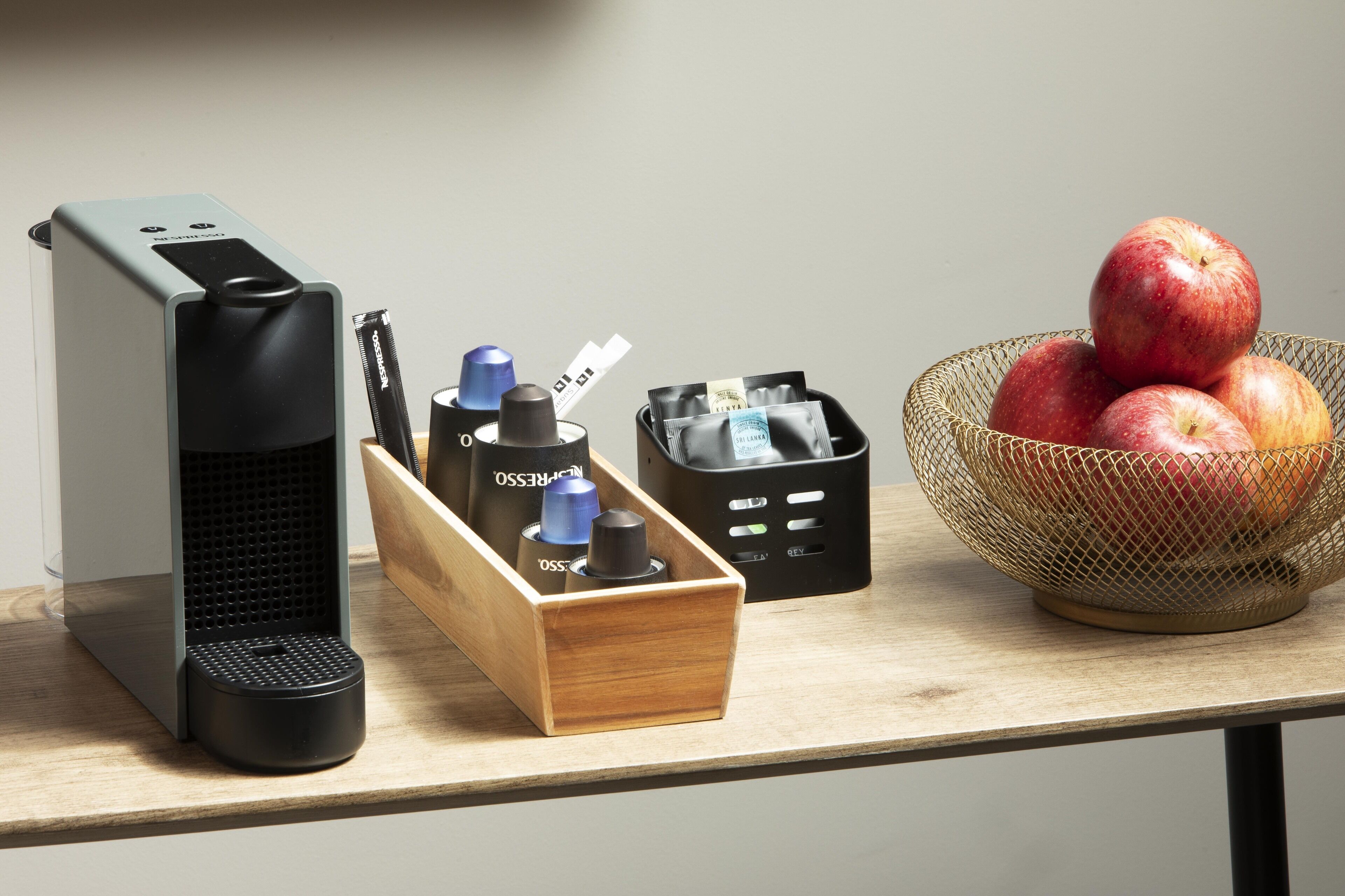 Un bureau avec une machine à café, une boîte en bois contenant des stylos, un récipient noir rempli de cartes et un panier de pommes.