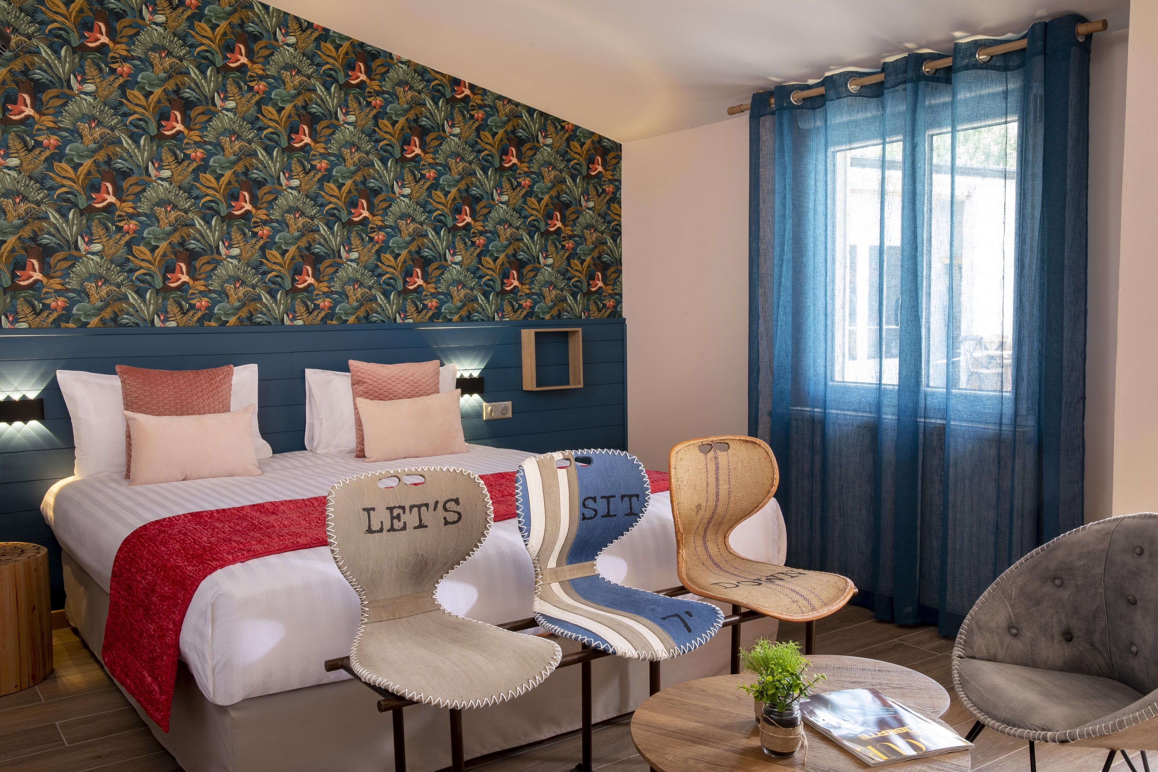 A hotel room with a patterned wallpaper, a bed with a red blanket, two upholstered chairs with 'LET'S SIT' signs, a small table with a plant, and a window with blue curtains.