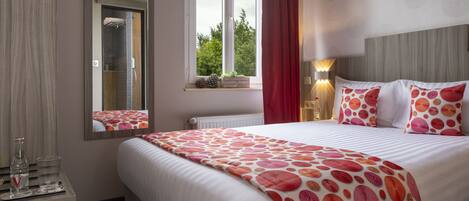 A hotel room with a large bed, a bedside table with glasses and a bottle, a window with a view of trees, and a red curtain.