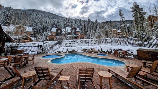 Un espace piscine doté de chaises longues et d’une terrasse en bois, entouré d’arbres et de bâtiments recouverts de neige.