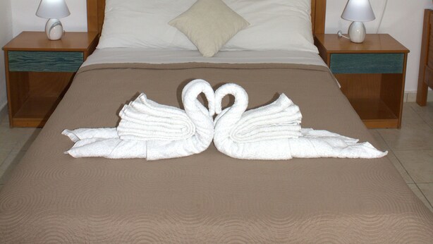 A neatly made bed with a swan towel design, wooden headboard, two bedside tables with lamps, and a painting above the bed. - Armonia Beach apartment 1 (Δήμος Κορισσίων)