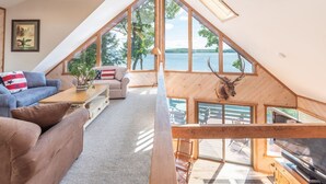 A loft-style room with a large window overlooking a lake, wooden beams, a ceiling fan, and a mounted deer head. - Stunning Waterfront Home with Boat Dock, swim, boating. Bike & skiing nearby. (Moultonborough)