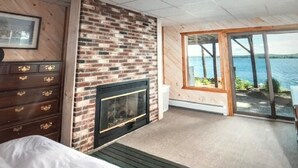 A room with a brick fireplace, wooden furniture, and a large sliding glass door leading to a balcony with a view of the water. - Stunning Waterfront Home with Boat Dock, swim, boating. Bike & skiing nearby. (Moultonborough)