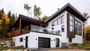 A modern two-story house with a large garage, a balcony, and a view of the surrounding forest. - The Arial cottage, spectacular view and Spa (La Malbaie)