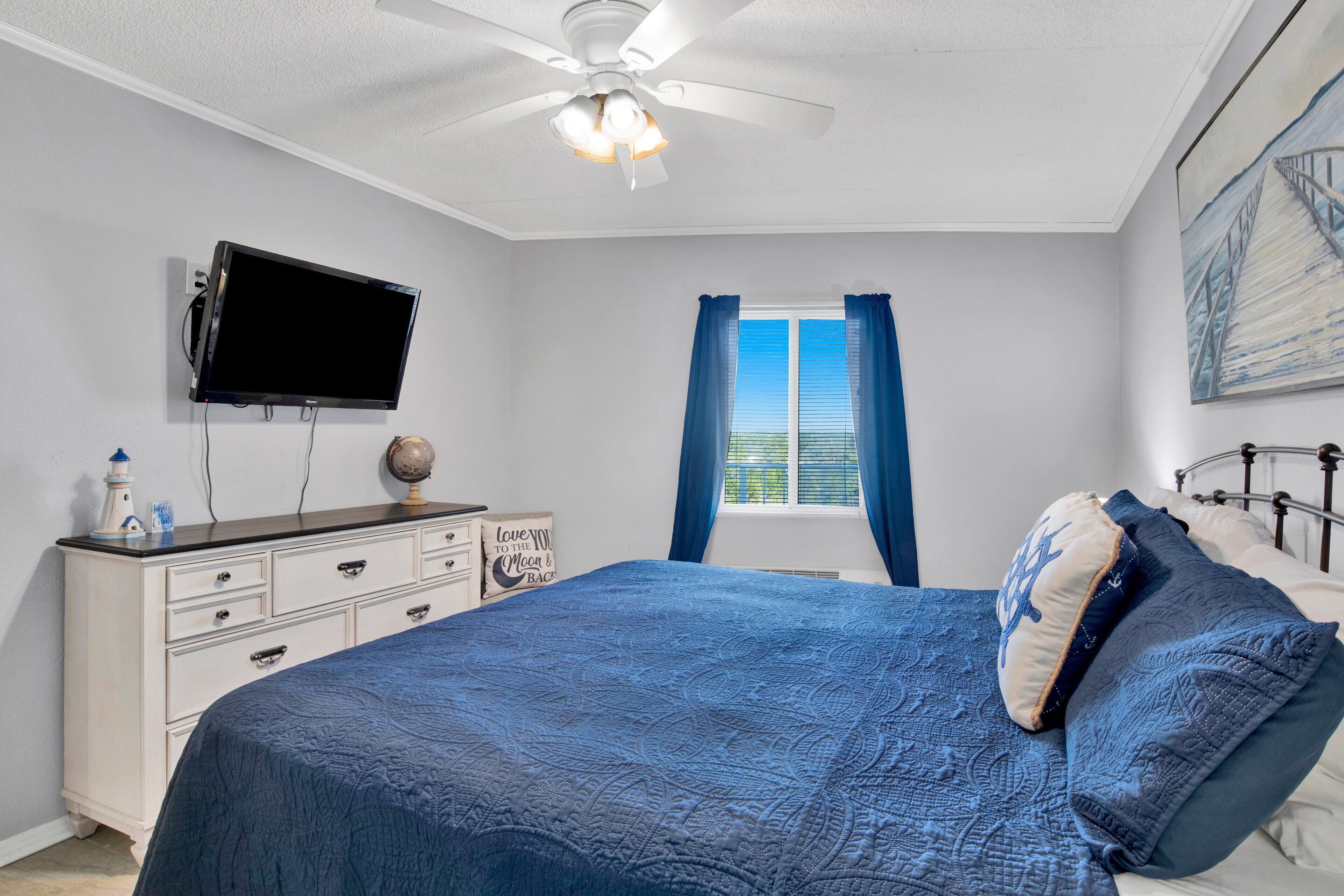 A bedroom with a bed, a ceiling fan, a TV mounted on the wall, a dresser, and a window with curtains.