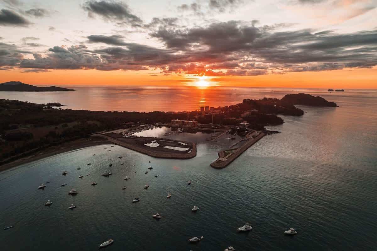Vista aérea de un puerto costero con barcos amarrados y un atardecer al fondo.