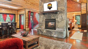 A cozy cabin interior with a stone fireplace, wooden walls, a dining area with chairs, and a large window with red curtains. - Love Me Smore in Hochatown/Broken Bow,OK (Broken Bow)