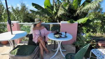 Une personne est assise à une table, avec une boisson et des en-cas, entourée de meubles en osier et de plantes en pot, dans un cadre extérieur ensoleillé.