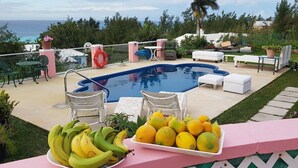 Zona de piscina con sillones de descanso, una mesa con fruta y vista al océano.