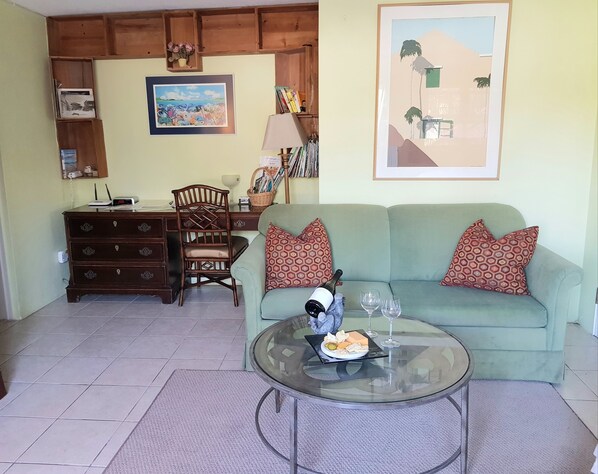 A living room with a green sofa, a glass coffee table with snacks and drinks, a wooden cabinet, a desk with a lamp, and a framed picture on the wall. - Bermuda Connections Guest House (Southampton Parish)