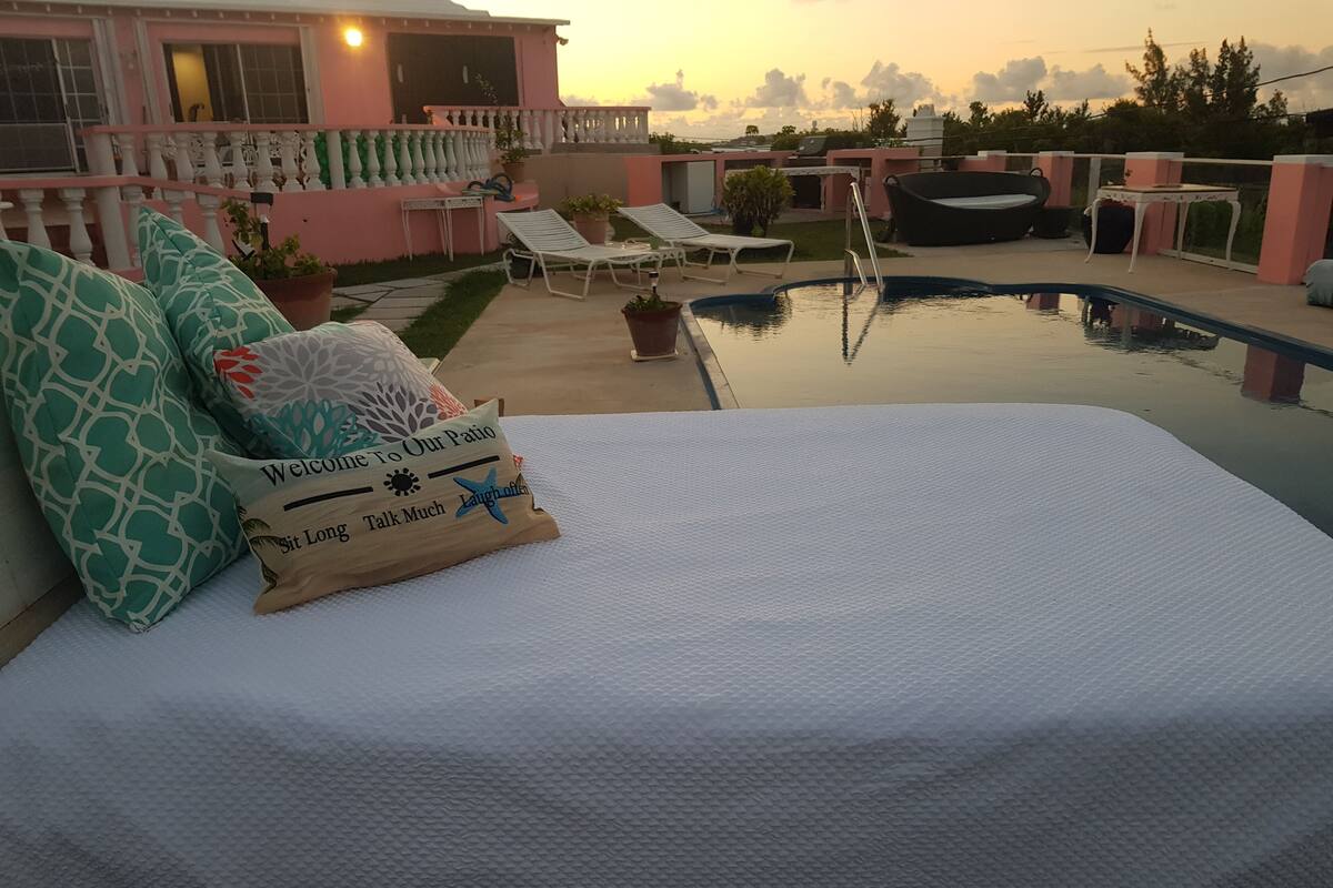 A bedroom with a pool view, a bed with a pillow, lounge chairs, and a pink building in the background.
