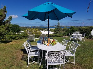 Un patio al aire libre con una mesa redonda y sillas de metal blanco, un paraguas azul y diversos alimentos.