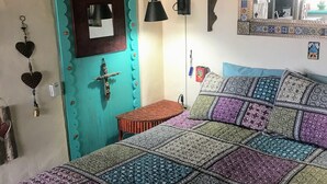 Een slaapkamer met een turquoise deur, een bed met een kleurrijke quilt, een wandlamp en een decoratieve spiegel.
