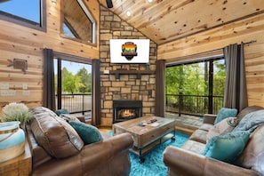 A cozy cabin interior with a stone fireplace, leather sofas, and large windows overlooking a forest.