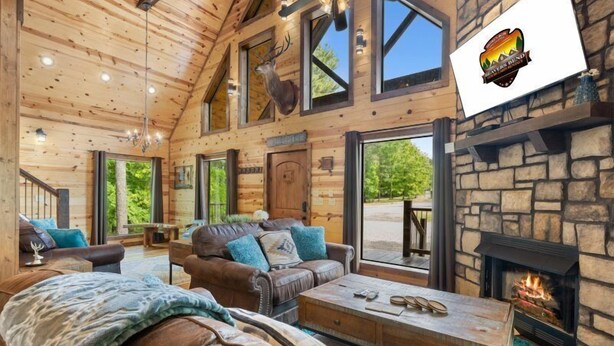 Ein Blockhaus-Interieur mit Kamin aus Stein, Ledersofas und einem großen Fenster mit Blick auf Grünflächen.