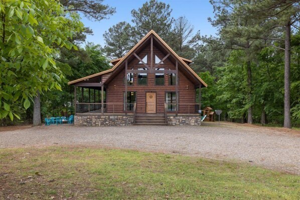 A wooden cabin with a stone foundation, a porch with chairs, and a gravel driveway. - Dreams Come True in Hochatown/Broken Bow, OK! (Broken Bow)
