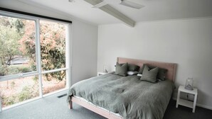 Ein Schlafzimmer mit großem Fenster, einem Bett mit grauer Bettwäsche, zwei Nachttischen und Blick auf Bäume.