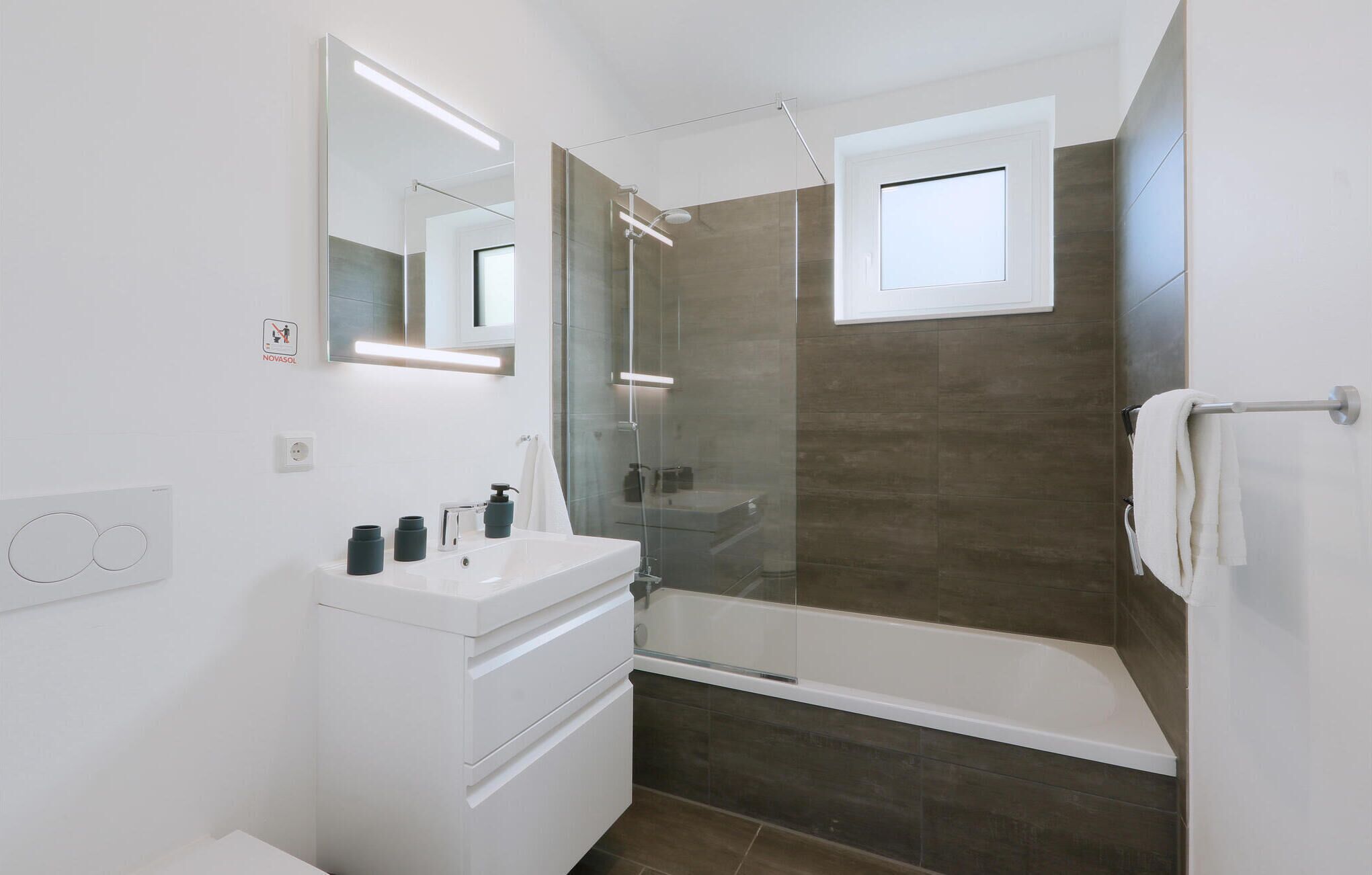 A modern bathroom with a glass-enclosed shower, a white vanity with a mirror, and a small window above the shower.