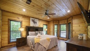 Ein Schlafzimmer in einer Holzhütte mit Bett, Nachttischen, Deckenventilator und Fenster mit Blick auf Bäume.