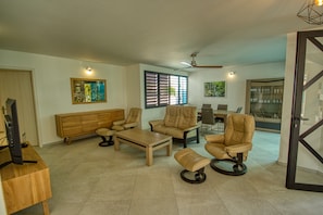 Uma sala de estar ampla com um sofá, duas poltronas, uma mesa de centro e uma TV sobre uma mesa de madeira. Há um ventilador de teto, uma janela com persianas e um aparador de vidro com objetos decorativos.