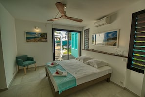 Quarto com cama, ventoinha no teto, cadeira e vista para o exterior.