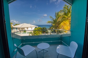 Varanda com duas cadeiras brancas e uma mesa pequena, com vista para uma praia com palmeiras e um edifício branco.