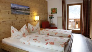 A double bed with floral bedding, a wooden headboard, a nightstand with a lamp, a red chair, and a window with curtains. - Pederü (Marebbe)