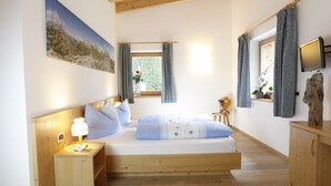A bedroom with a bed, bedside table, lamp, window with curtains, and a mounted TV. - Pederü (Marebbe)