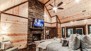 Interior de una cabaña de troncos con chimenea de piedra, un televisor montado encima y una cama con muchas almohadas.