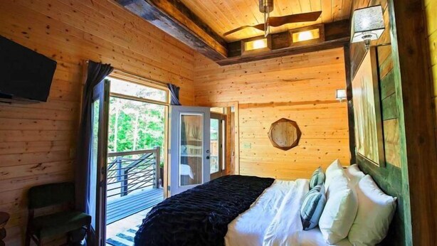 A wooden cabin bedroom with a bed, a ceiling fan, a TV, and a balcony door.
