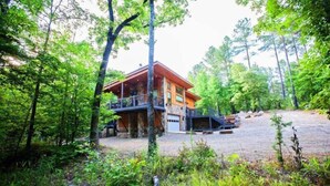 A cabin with a wooden exterior, a balcony, and a stone foundation, surrounded by dense forest.