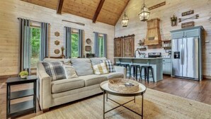 Ein gemütliches Blockhaus-Interieur mit Holzbalkendecke, einer Couch mit Kissen, einem Essbereich und einer Küche mit Kühlschrank und Barhocker.