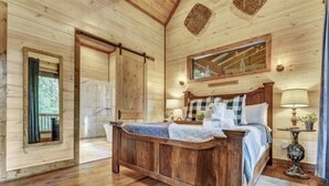Ein Schlafzimmer einer Holzhütte mit einem großen Bett, einem Fenster mit Vorhängen, einer Holzbalkendecke und einem kleinen Tisch mit Lampe.