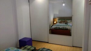 Een slaapkamer met een bed, een klein paars voetenbankje en een spiegel die de kamer weerspiegelt.