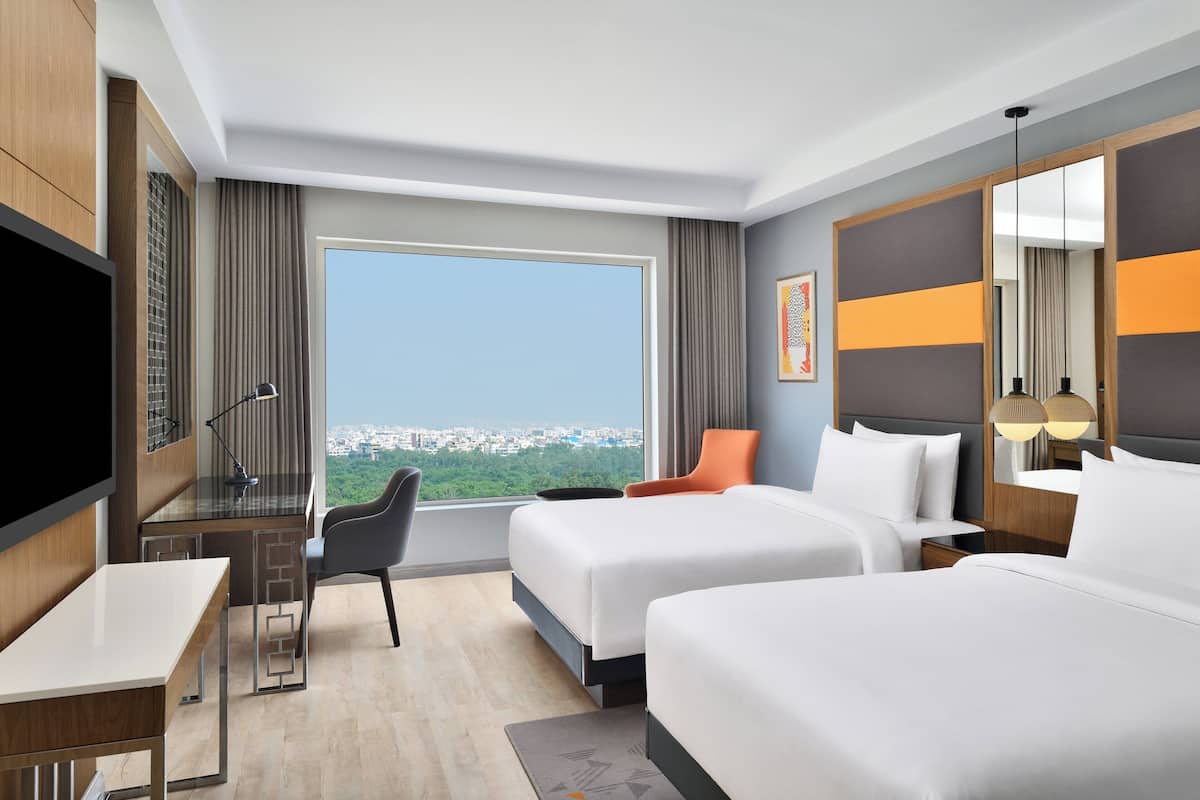 Ein Hotelzimmer mit zwei Betten, einem Schreibtisch, einem Sessel, einem Fernseher und einem großen Fenster mit Blick auf die Stadt.