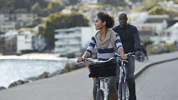 A person riding a bicycle on a paved path alongside a body of water, with buildings in the background.