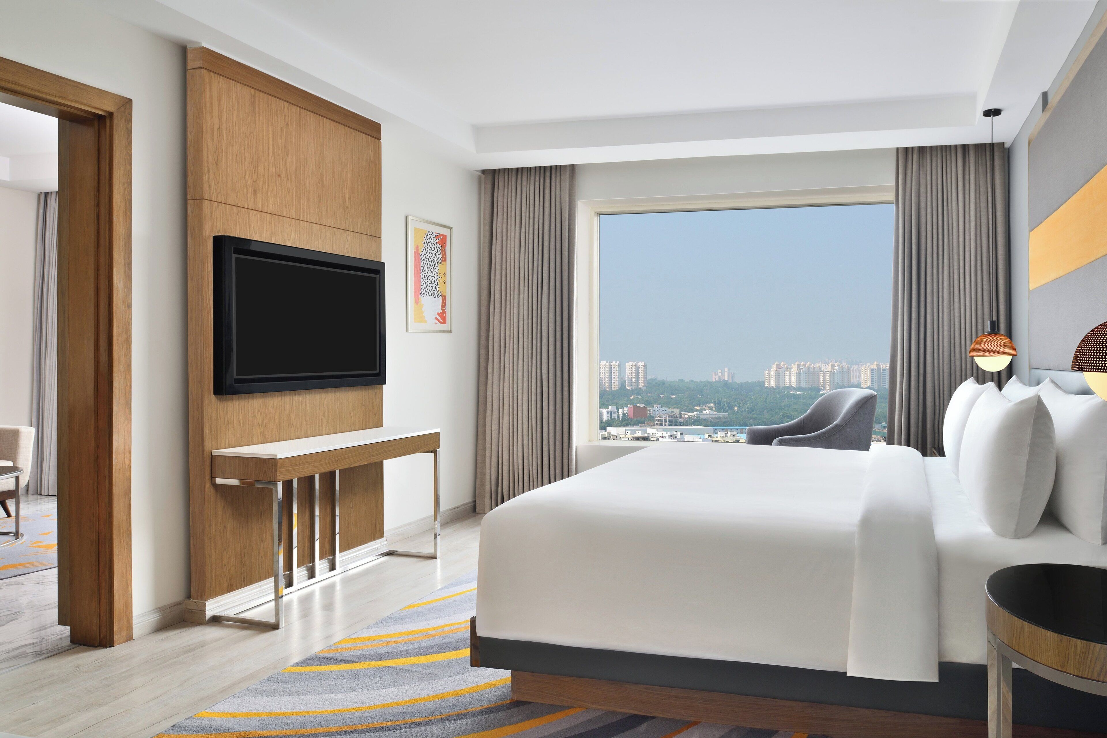Una habitación de hotel moderna con una cama grande, un televisor de pantalla plana en la pared, un escritorio y vistas a la ciudad a través de un ventanal amplio.