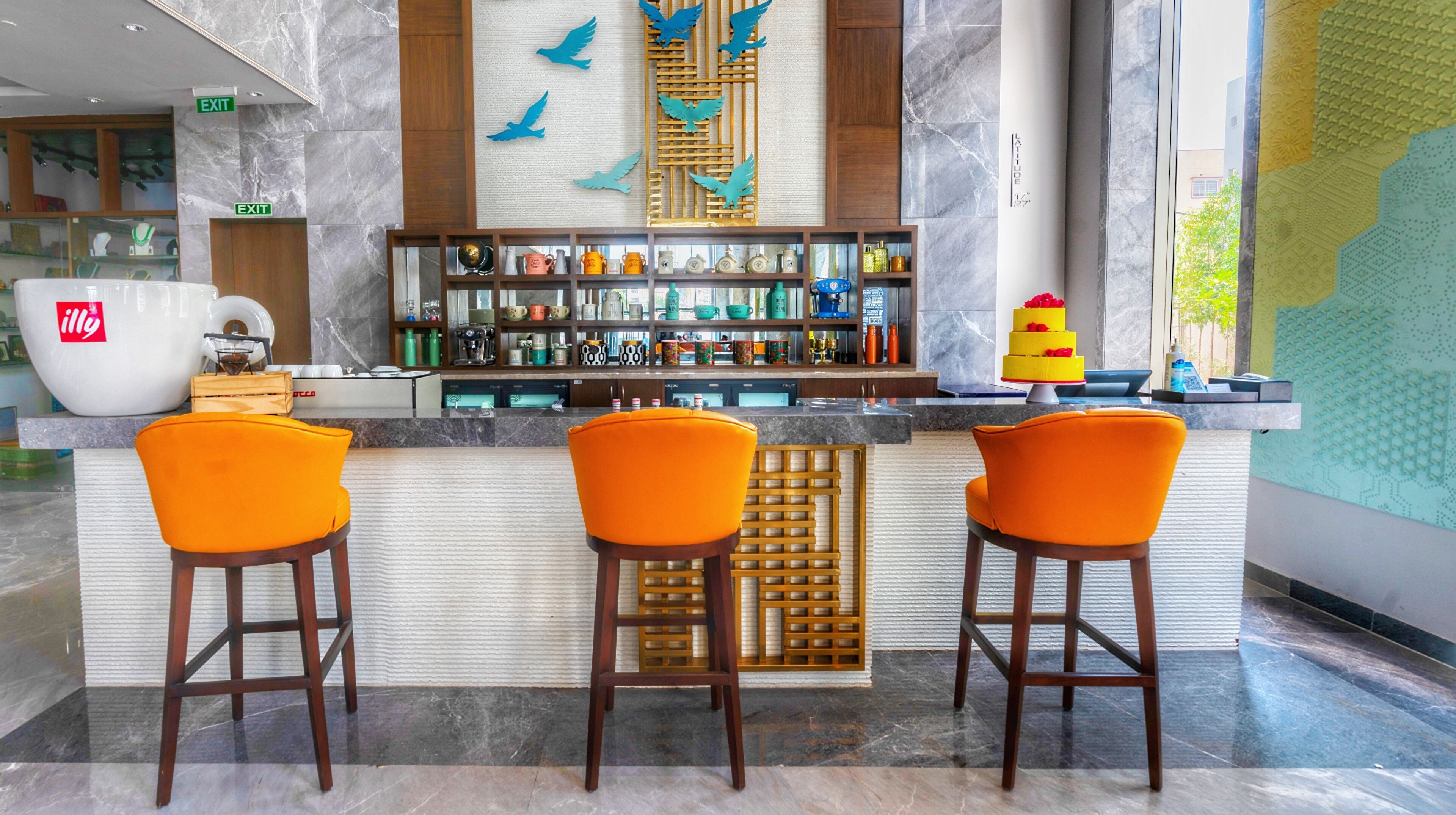 Zona de bar con tres sillas naranjas, un separador de madera y varios botellas y vasos en estantes.