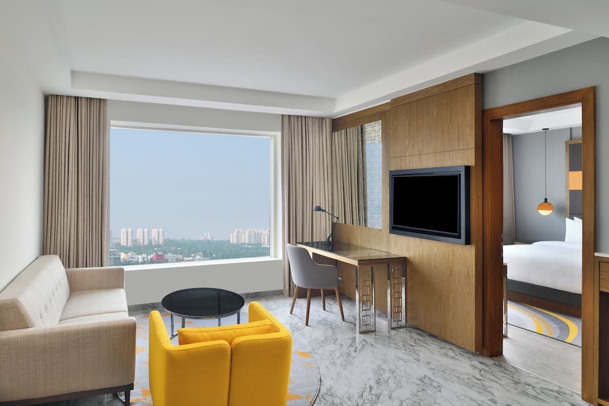 Ein Hotelzimmer mit einer Couch, einem gelben Sessel, einem Schreibtisch mit Stuhl, einem Fernseher und einem großen Fenster mit Blick auf die Stadt.