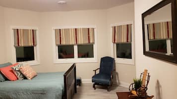 A bedroom with a bed, a chair, a small table, and three windows with curtains.