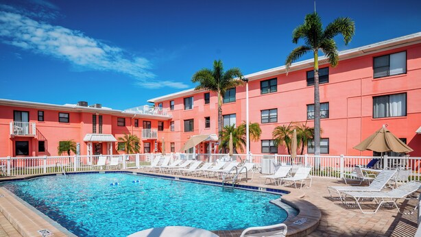 Piscina com espreguiçadeiras e guarda-sóis, cercada por um muro branco, com um edifício ao fundo.