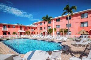 A swimming pool with lounge chairs and umbrellas, surrounded by a white fence, with a building in the background.